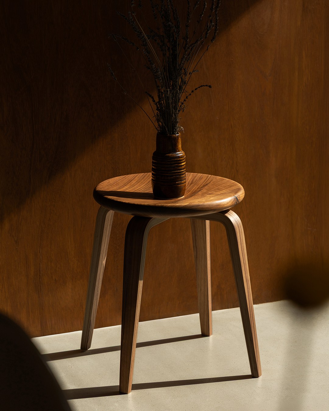 A Bistro stool with a carved walnut seat, holding a ribbed vase of dried grasses against a wood-panelled wall in late sunlight.
