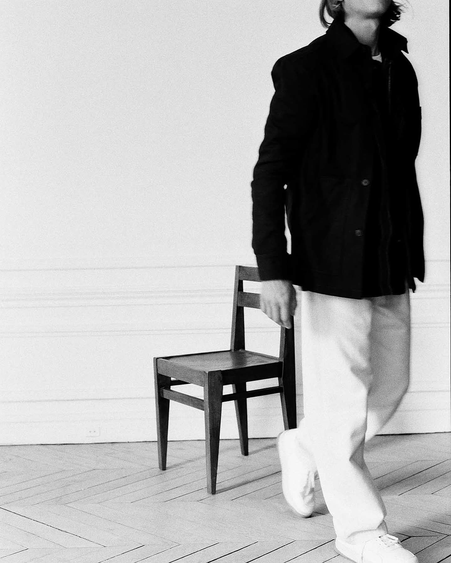 Model in a dark jacket and white trousers beside a chair for Haussmann by Zara.