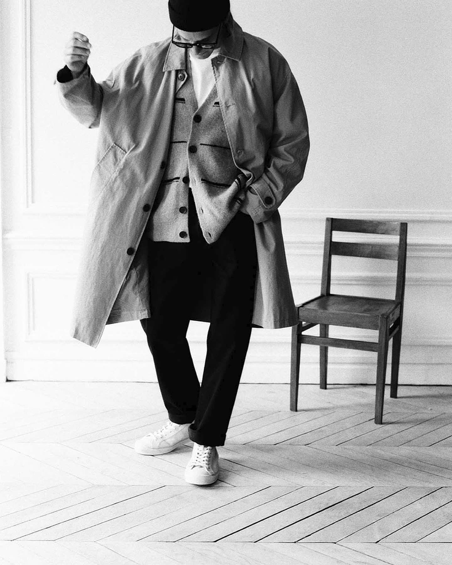 Model in a layered coat standing beside a wooden chair in a Parisian apartment for Haussmann by Zara.