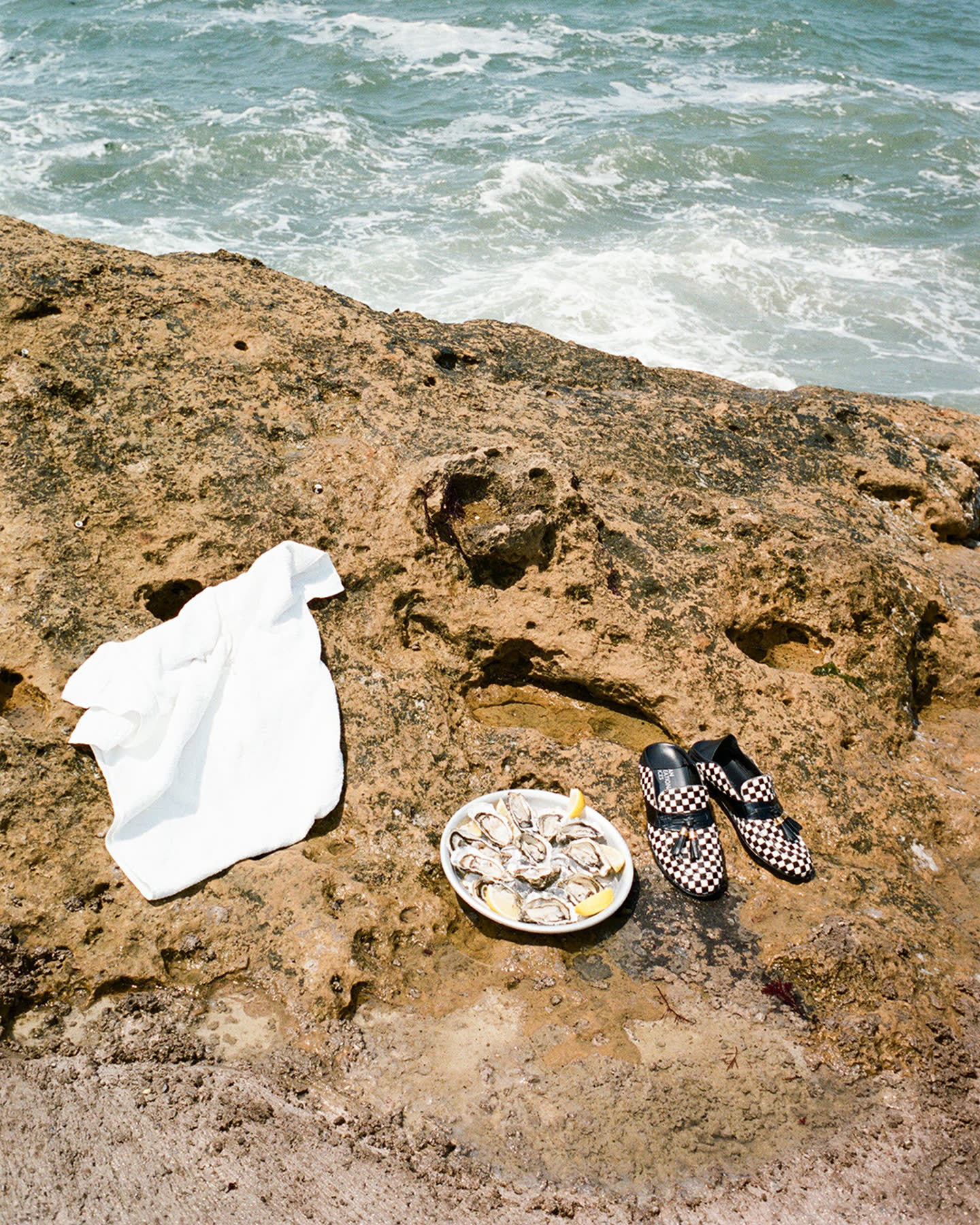 Oysters, white cloth, and checkerboard loafers arranged on the rocky Biarritz coast.