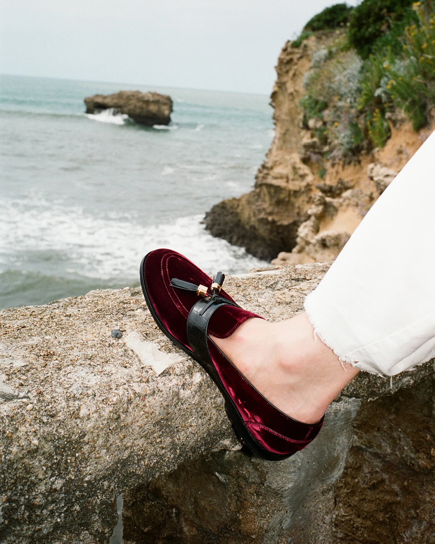 A burgundy loafer and white trouser leg above the rocky Biarritz coast.